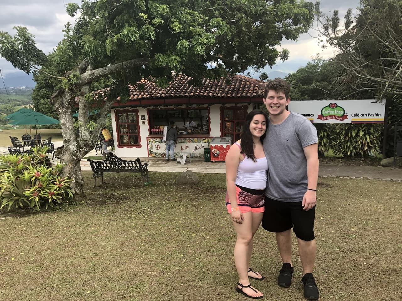 Sthephany and Seth in front of a small coffee shop under a big tree on a hillside in Colombia