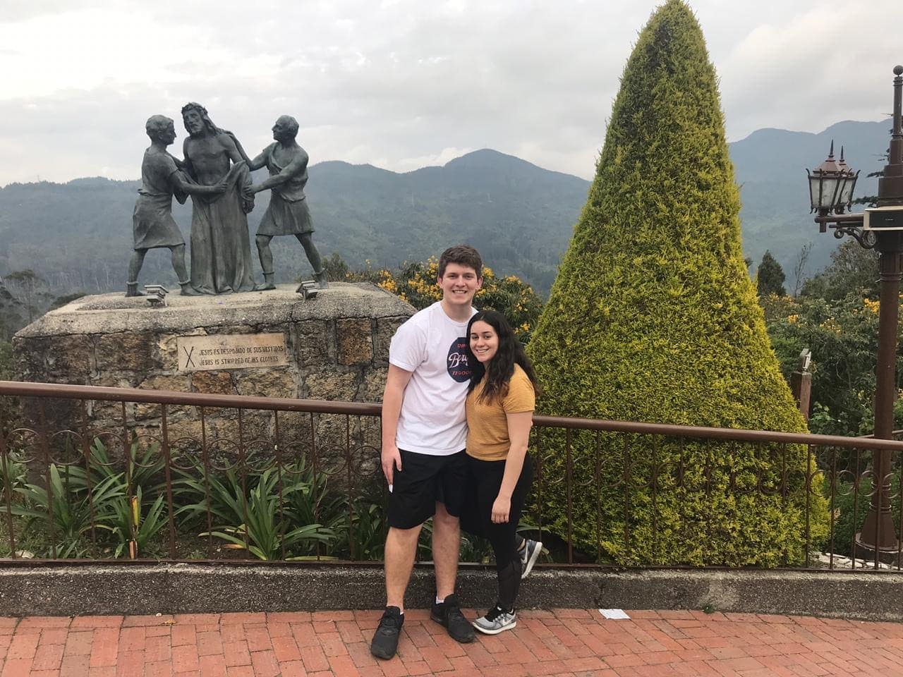 Sthephany and Seth posing together at a mountain viewpoint in Colombia, with statues, a trimmed cypress, and forested peaks behind them