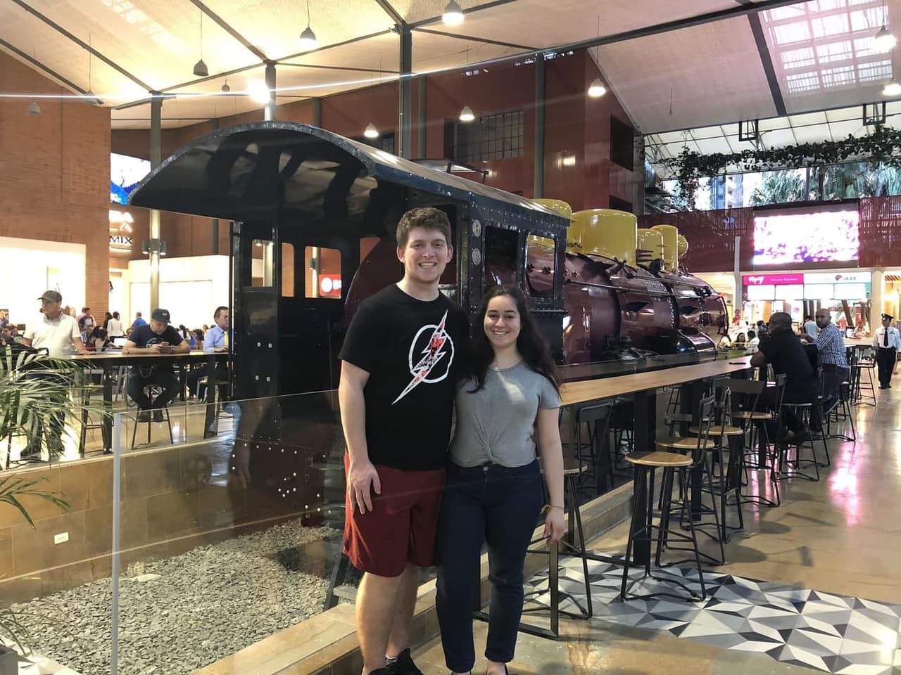Sthephany and Seth standing in front of a vintage steam locomotive displayed inside a shopping center in Colombia