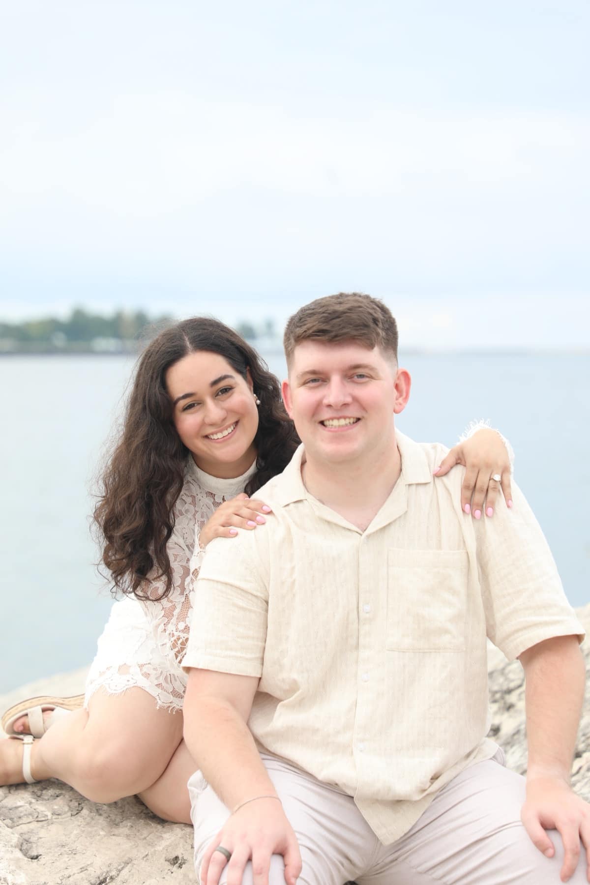 Sthephany and Seth smiling together on a rocky lakeshore, she in a white lace dress leaning on his shoulder, he in a cream linen shirt