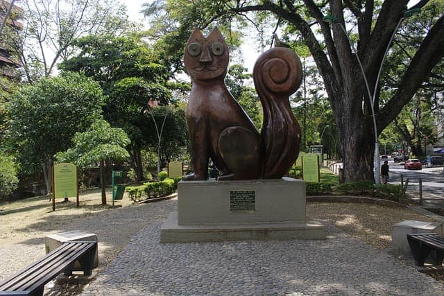 The large bronze cat sculpture, El Gato, standing on a stone base in a leafy park